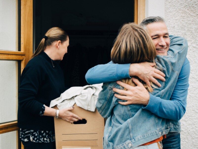 Photograph featuring a man hugging woman with a volunteer carrying a box into a house in the background