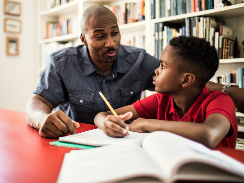 Man and middle-school aged son sitting at a red table working through homework using a textbook and notebook with a large shelf of books behind them.