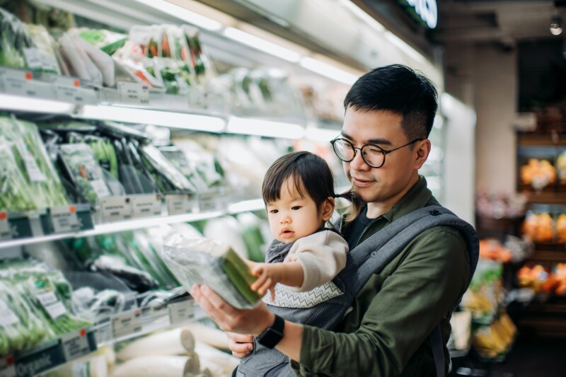 Young Asian father with baby grocery shopping for fresh vegetables in supermarket