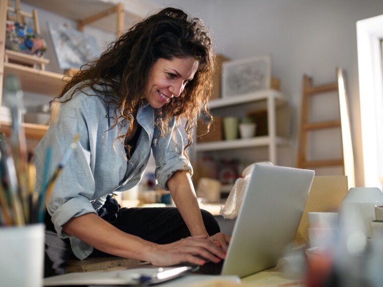 Happy young woman with laptop handles orders in pottery workshop.