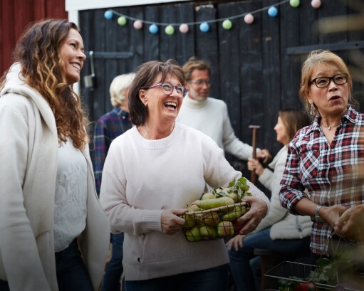 Three women smiling at an outdoor party, one holds a basket of root vegetables