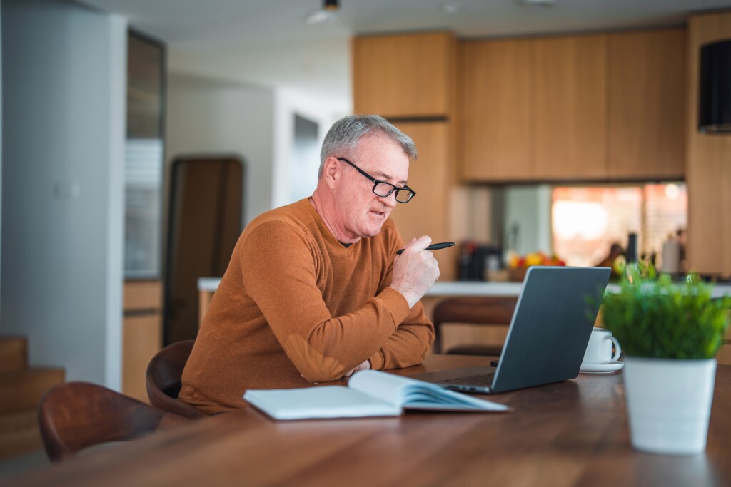 Mature Caucasian male with gray hair and eyeglasses sitting in a domestic dining room using laptop and taking notes.