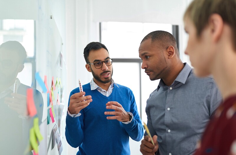 A group of workers are standing next to a whiteboard while one person leads the discussion