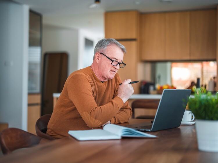 Mature Caucasian male with gray hair and eyeglasses sitting in a domestic dining room using laptop and taking notes.