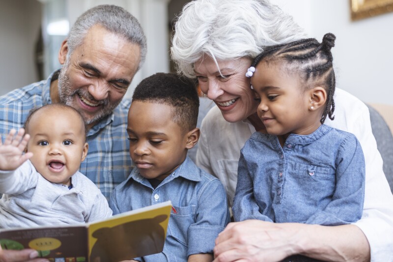 A retired senior couple support family by babysitting. Three young children sit on their grandparents' laps. The group is reading a book together.