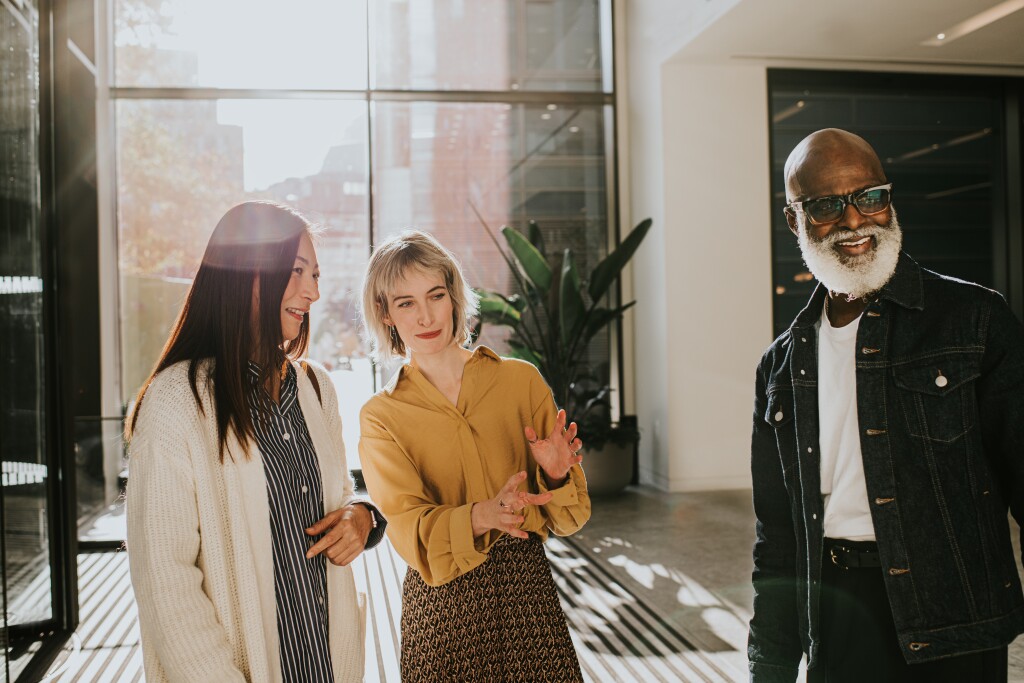 Three people stand in a group in a sunny office reception. One of woman explains something to the other two participants.