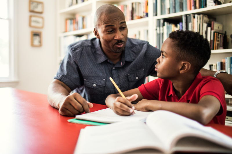 Man and middle-school aged son sitting at a red table working through homework using a textbook and notebook with a large shelf of books behind them.