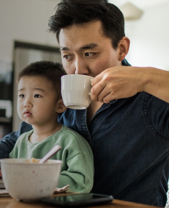 Toddler son sits in dad's lap while working on his laptop at the kitchen table eating breakfast