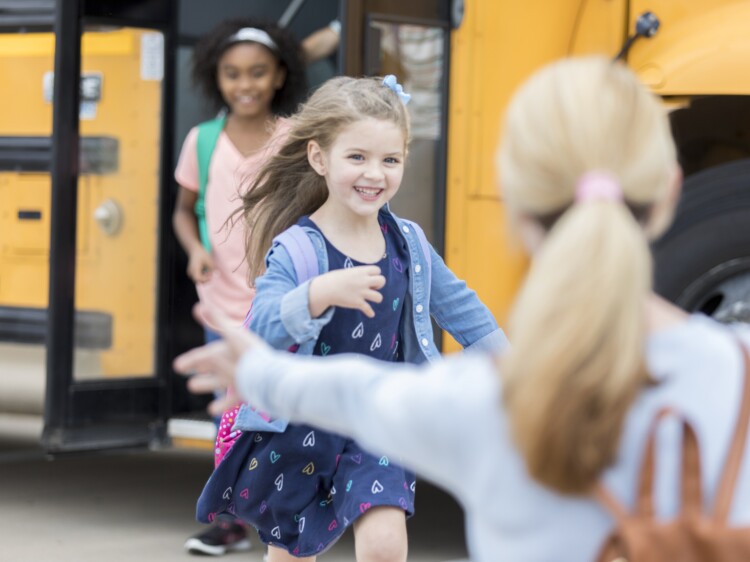 Excited little girl runs to her mom after getting off of the school bus. Her mom is waiting with her arms open wide.