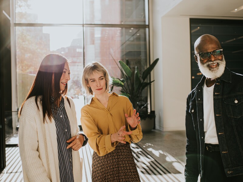 Three people stand in a group in a sunny office reception. One of woman explains something to the other two participants.
