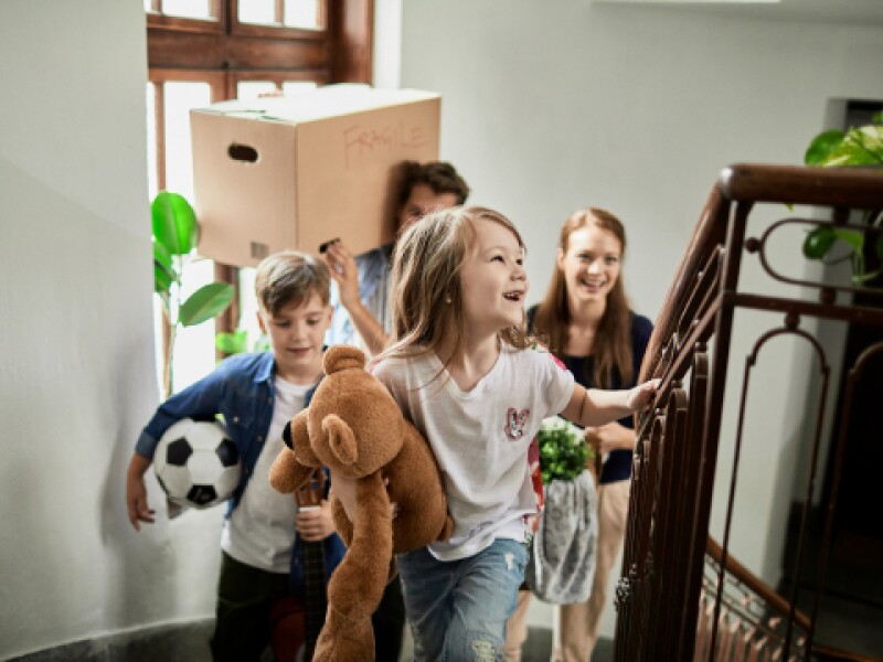 Family with two young children climb the stairs of a house with moving supplies, soccer ball, and teddy bear
