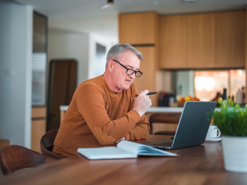 Mature Caucasian male with gray hair and eyeglasses sitting in a domestic dining room using laptop and taking notes.